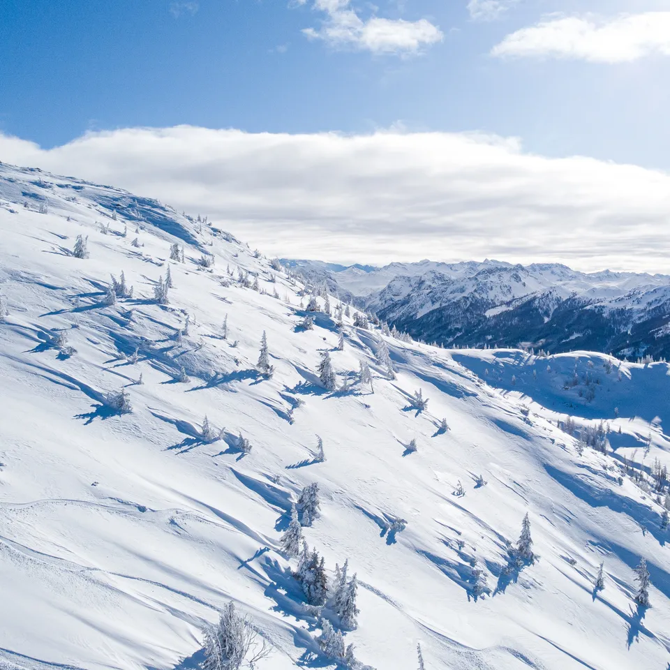 Verschneiter Berghang mit Bäumen unter blauem Himmel mit Wolken