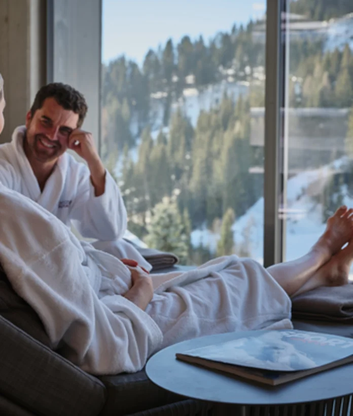 Paar in Bademänteln entspannt vor Panoramafenster mit Blick auf verschneite Berge