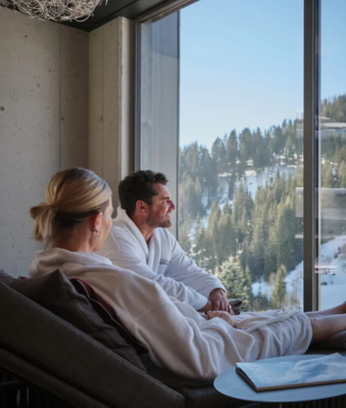 Paar in Bademänteln entspannt vor Fenster mit Blick auf verschneite Landschaft
