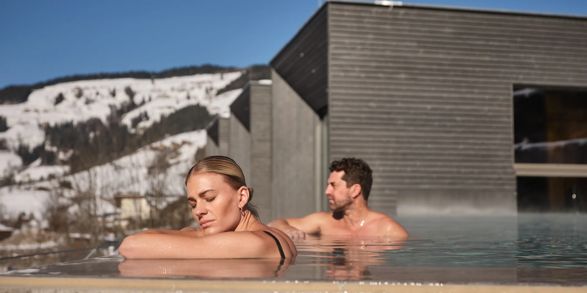Woman and man relaxing in the outdoor pool of Sporthotel Wagrain  in front of snow-capped mountains