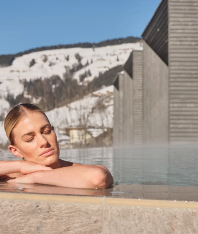 Frau entspannt im Außenpool vor schneebedeckten Bergen und Hotelgebäude