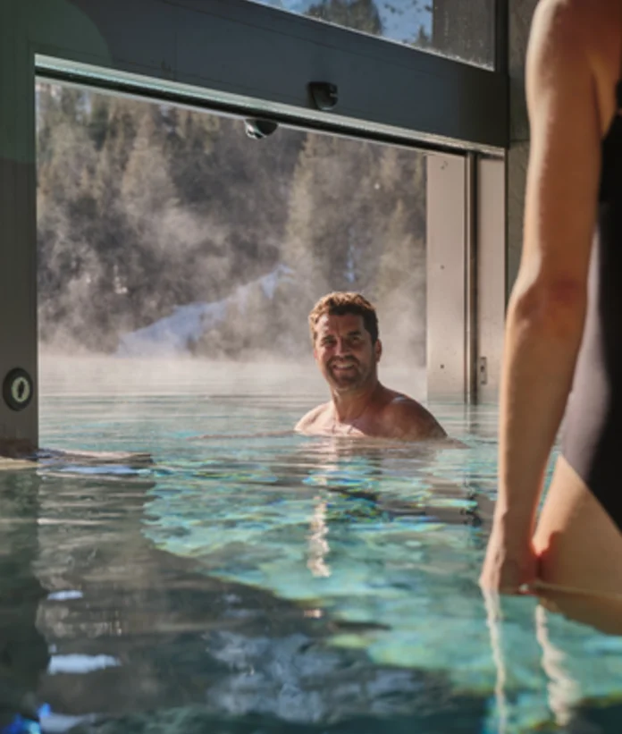 Man in pool, woman in foreground, snowy mountains in background