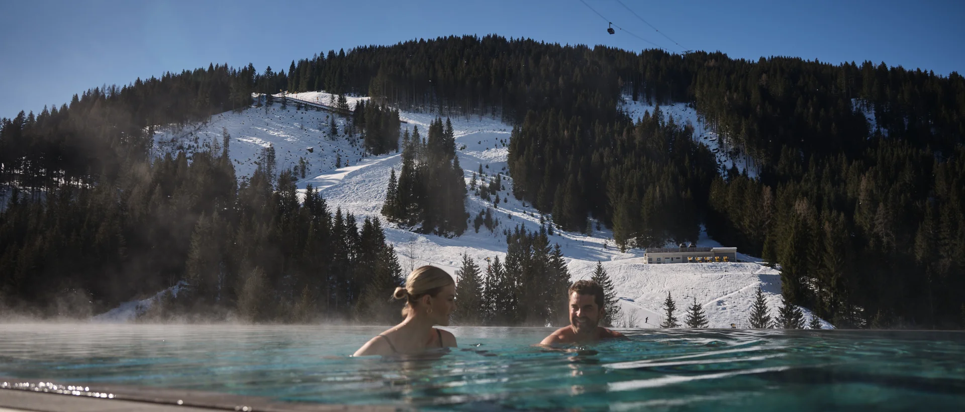 Two people in the outdoor pool in front of a snowy mountain landscape