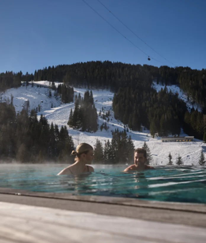 Two people in the outdoor pool in front of a snowy mountain landscape