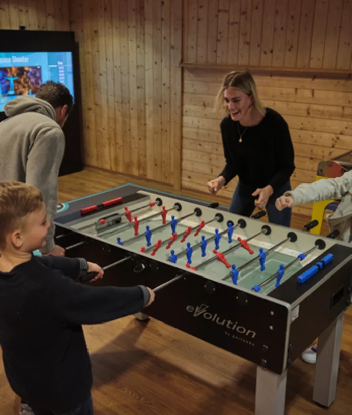 Familie spielt Tischfußball in einem Raum mit Holzvertäfelung und einem Bildschirm