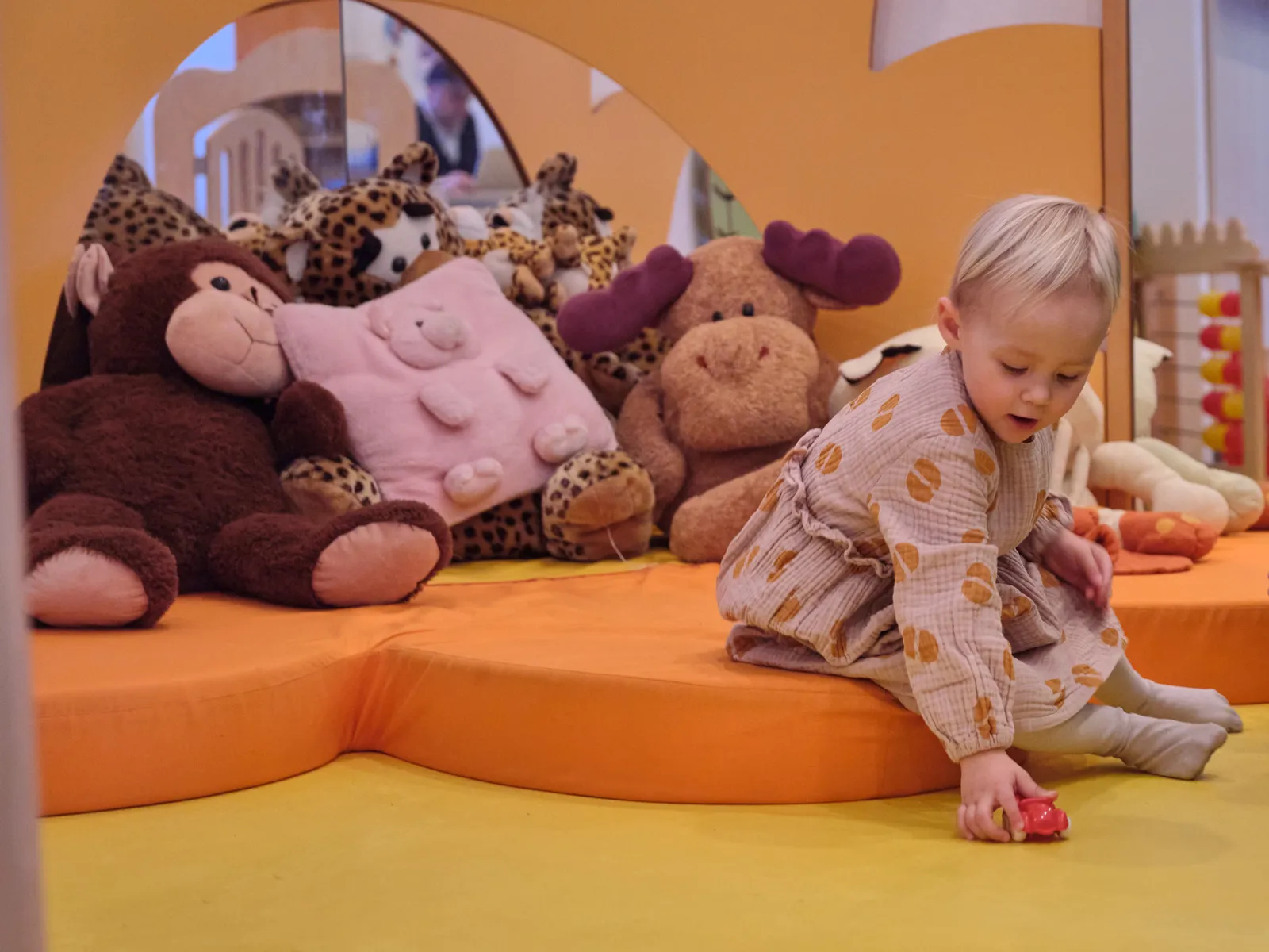 Kleines Mädchen spielt auf orangefarbener Fläche mit Stofftieren im Hintergrund