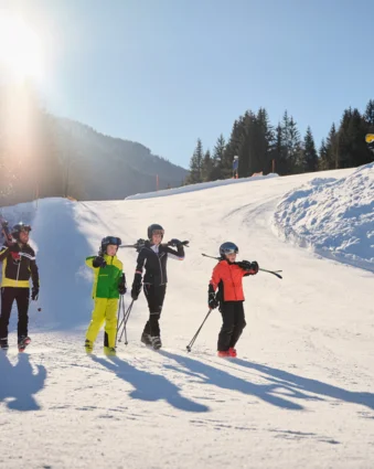 Familie mit Skiern und Stöcken auf schneebedeckter Piste vor Bergkulisse